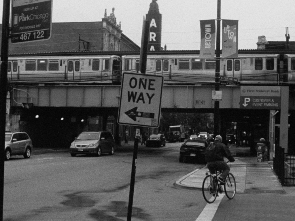 Black and white street photograph featuring 'ONE WAY' road sign, a passing train over a bridge, someone riding a bike and parked cars